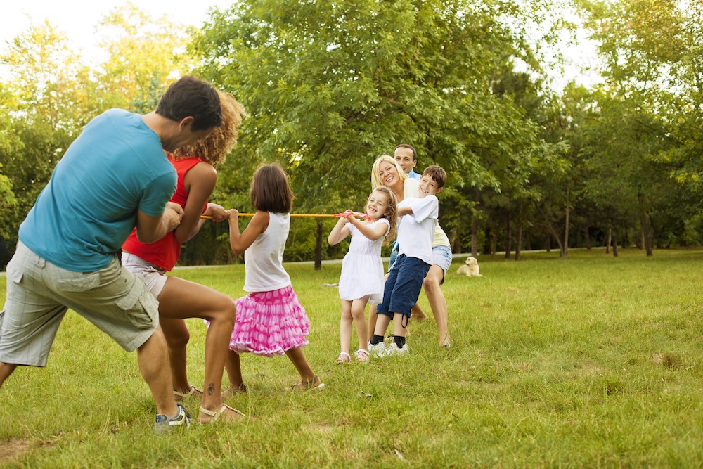 People, including children and adults, playing tug of war on a grassy field in a park with trees in the background.