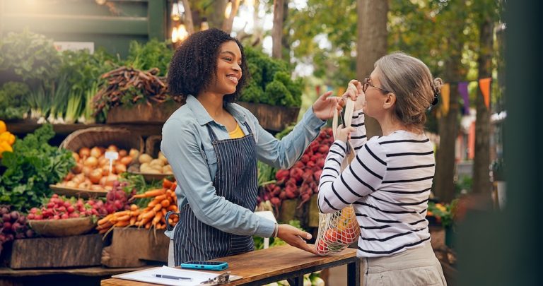 A vendor and customer smile while exchanging produce at an outdoor farmers market stand full of fresh vegetables.
