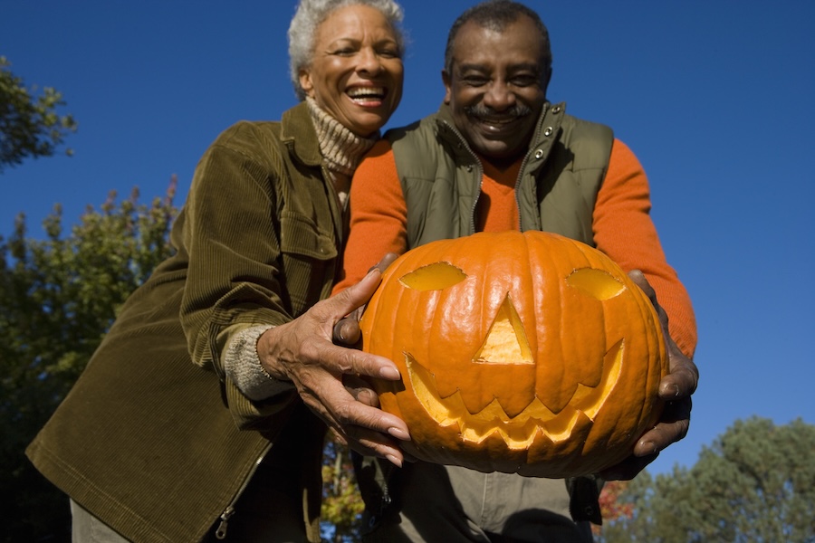 Smiling couple outdoors holding a carved jack-o'-lantern pumpkin on a sunny day.