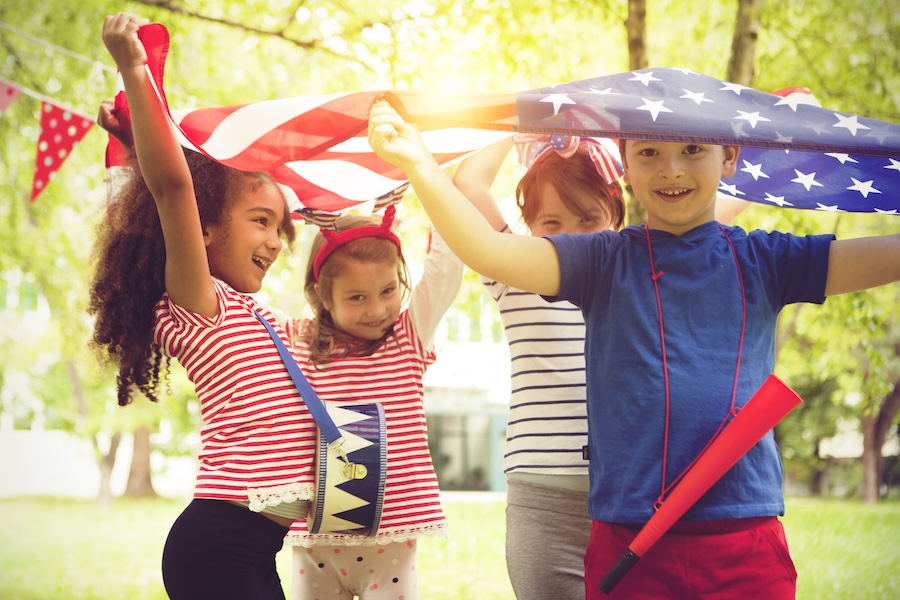 Four children celebrate outdoors with American flags and patriotic outfits, smiling and having fun together.