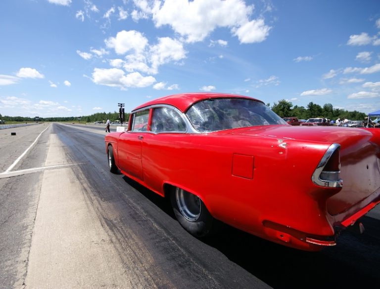 A bright red vintage car on a drag strip, ready to race under a sunny sky with scattered clouds.