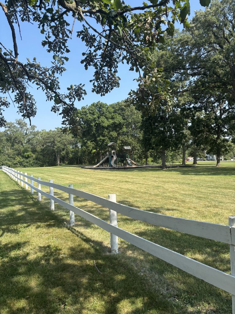 A playground with a slide and swings sits beyond a white fence in a grassy, tree-lined park.