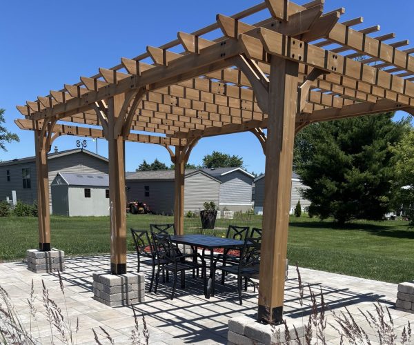 Wooden pergola over a patio table and chairs, set on stone tiles with grass and houses in the background.