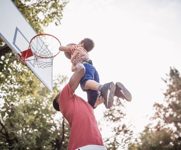 An adult lifts a child towards a basketball hoop outdoors at Glenwood Manufactured Home Community, helping him dunk the ball on a sunny day.